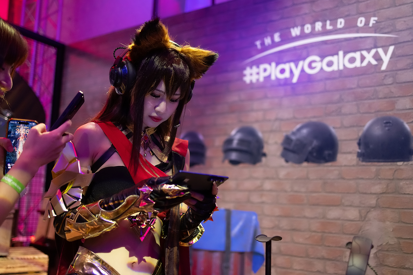 A woman wearing a cosplay costume and a headset stares at her mobile phone in front of a wall with The World of PlayGalaxy branding.