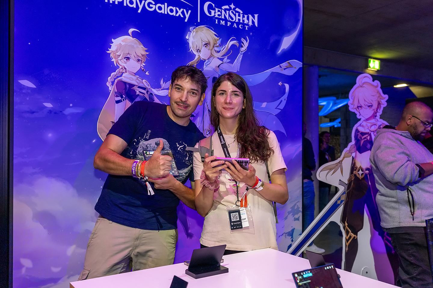 A man and a woman stand in front of the Genshin Impact game banner at The World of PlayGalaxy booth, facing the camera.