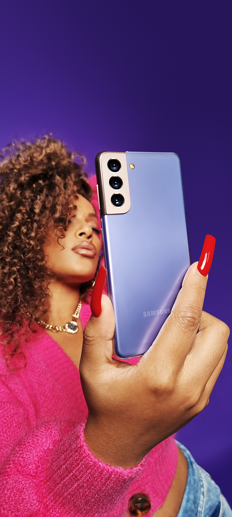 a photo captured with a galaxy s21 ultra showing a woman in pink cardigan taking a selfie with a violet galaxy s21 plus against a purple backdrop
